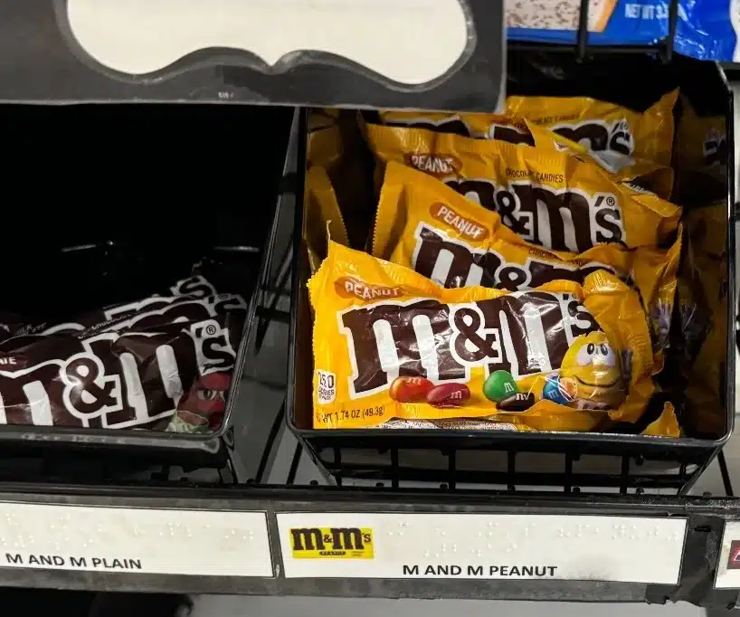 Basket of individual M&M’s bags with braille and text sign describing product.